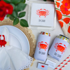Table setting with 'OH CRAB!' themed can coolers, napkins, and straws on a white surface.