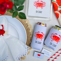 Cans with 'OH CRAB!' can coolers  on a table setting with napkins, matchboxes and straws.