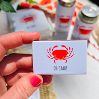 Hand holding a matchbox with a red crab design and 'OH CRAB!' text, with drinks and straws in the background.