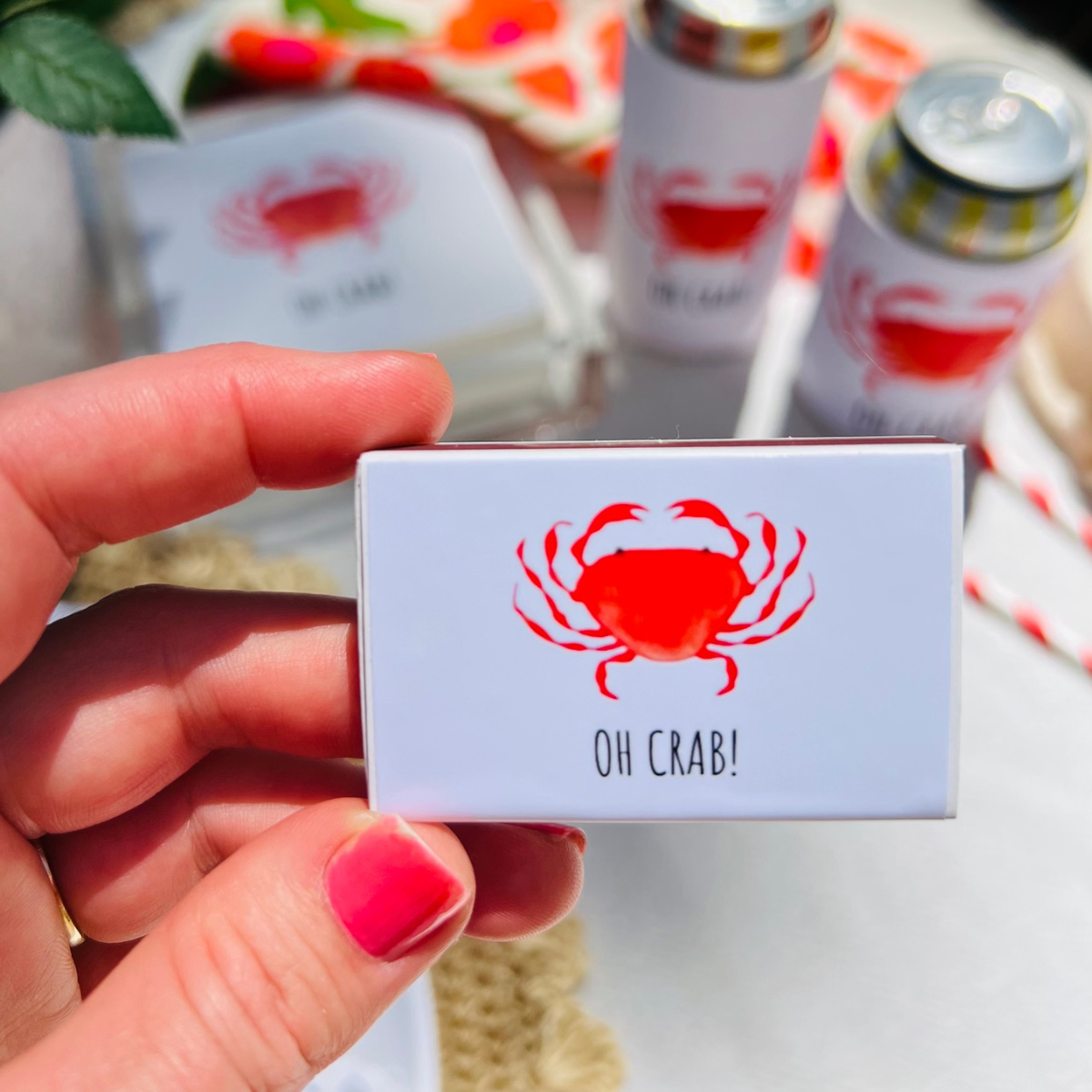 Hand holding a matchbox with a red crab design and 'OH CRAB!' text, with drinks and straws in the background.