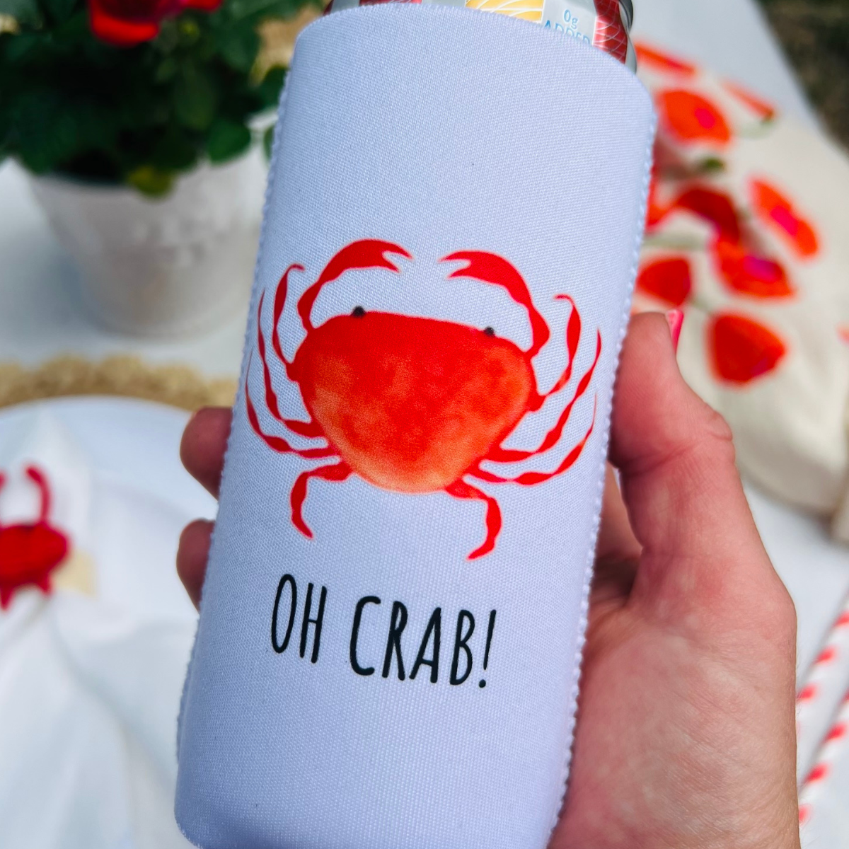 Hand holding a white can cooler with a red crab design and 'OH CRAB!' text, outdoor setting.