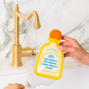 A hand holds a yellow and orange novelty tequila bottle-shaped sponge over a running faucet, with a glass below. The object has printed text reading 'One Tequila, Two Tequila, Three Tequila, Chore."