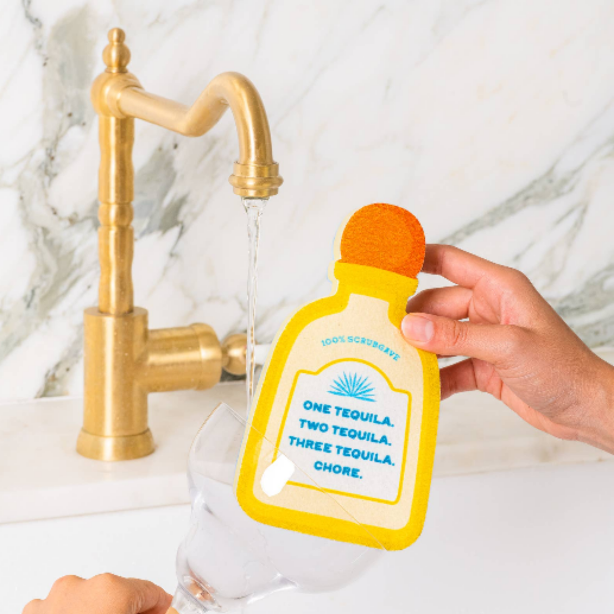 A hand holds a yellow and orange novelty tequila bottle-shaped sponge over a running faucet, with a glass below. The object has printed text reading 'One Tequila, Two Tequila, Three Tequila, Chore."