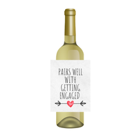 White wine bottle with a customizable engagement wine label is shown against a white background. The wine label has text that reads "Pairs well with an engagement" above a watercolor illustration of a red heart with leaves.