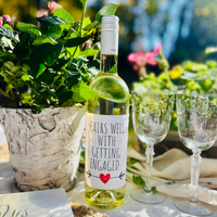 Bottle of white wine is shown on a table outdoors beside two glasses. The wine bottle has a fun engagement wine label that reads "Pairs Well With An Engagement."
