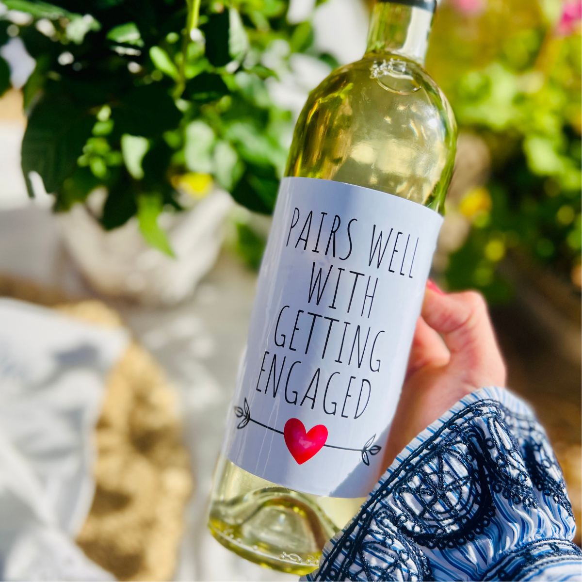 Bottle of white wine with an engagement wine label held by a person outdoors. The wine label reads "Pairs well with an engagement," and makes a fun engagement gift. 
