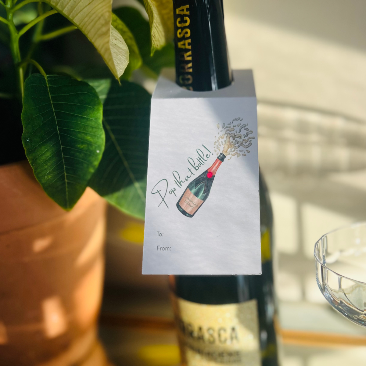 Bottle of champagne is pictures with a decorative wine label next to a potted plant. The wine label reads "Pop that bottle!" and features a champagne bottle with champagne bursting from the top. 