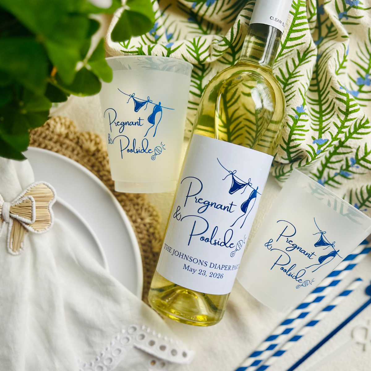 Bottle of white wine with 'Pregnant & Poolside' labels on a decorative tablecloth is shown beside two frost flex cups with a matching design for poolside summer baby showers. 