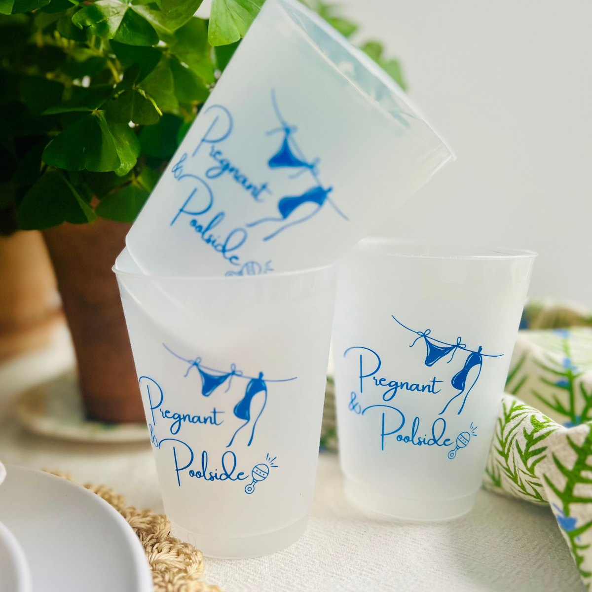 Frosted cups with 'Pregnant Poolside' design on a table with plants and a plant pot in the background.