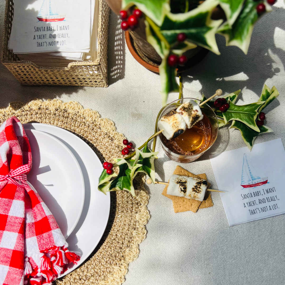 Santa Baby Holiday Napkins on table next to Christmas Cocktail styled with mistletoe, and Christmas place setting that features white plates styles with a gingham napkin that's tied with a bow