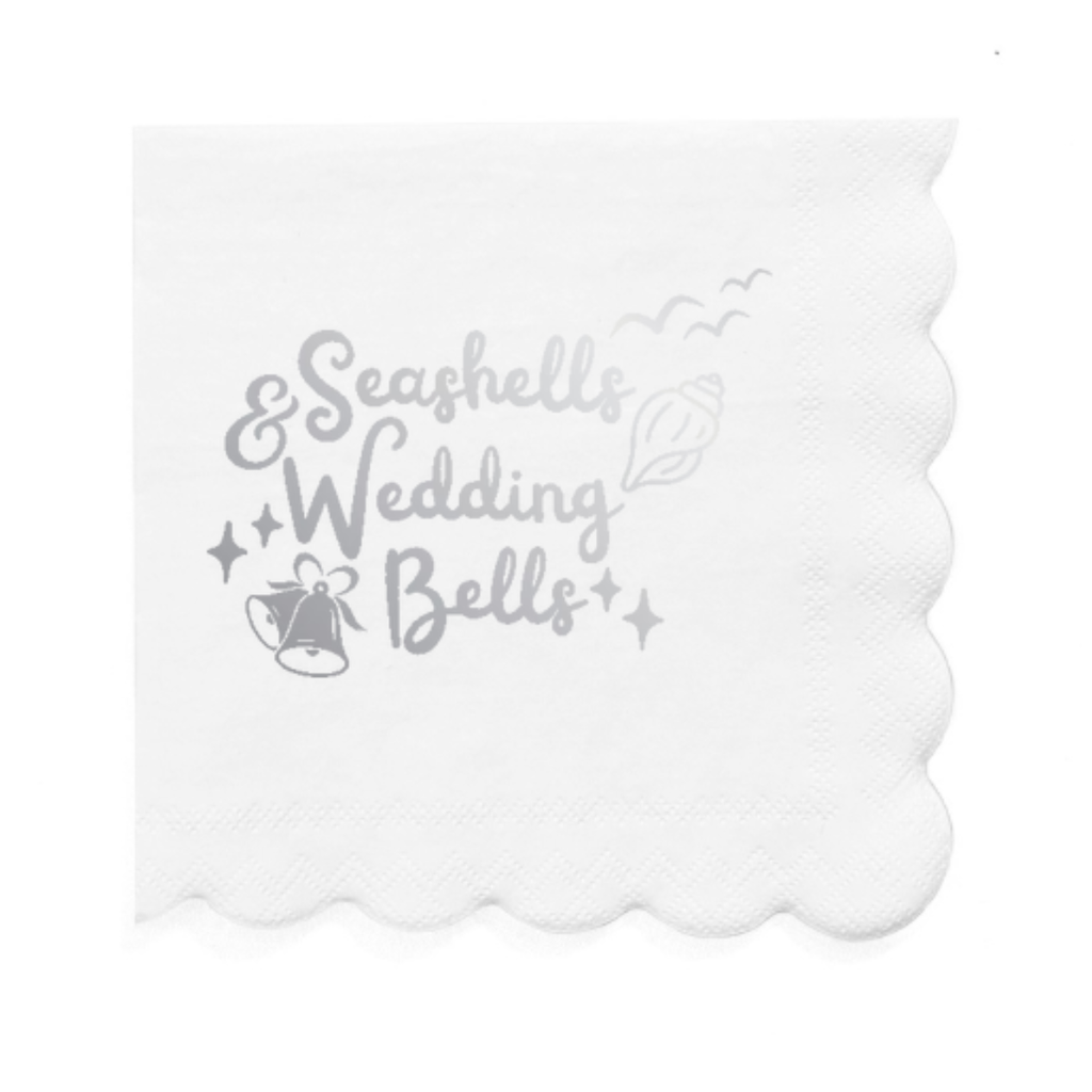 Decorative white paper cocktail napkin with 'Seashells & Wedding Bells' text and decorative elements in silver foil is shown on a white background.