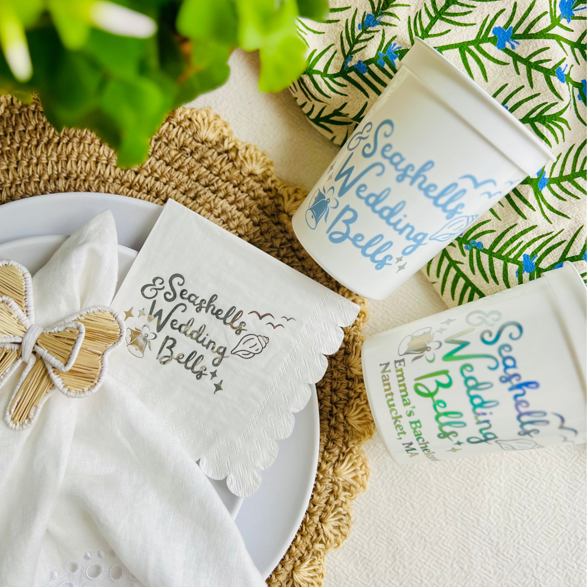 White paper scalloped cocktail napkin with silver foil text that reads "Seashells & Wedding Bells" is shown on a plate beside matching white stadium cups that feature the same design. 