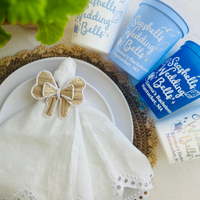 Napkin ring in the shape of a bow and 'Seashells Wedding Bells' customizable stadium cups on a table with decorative elements.  