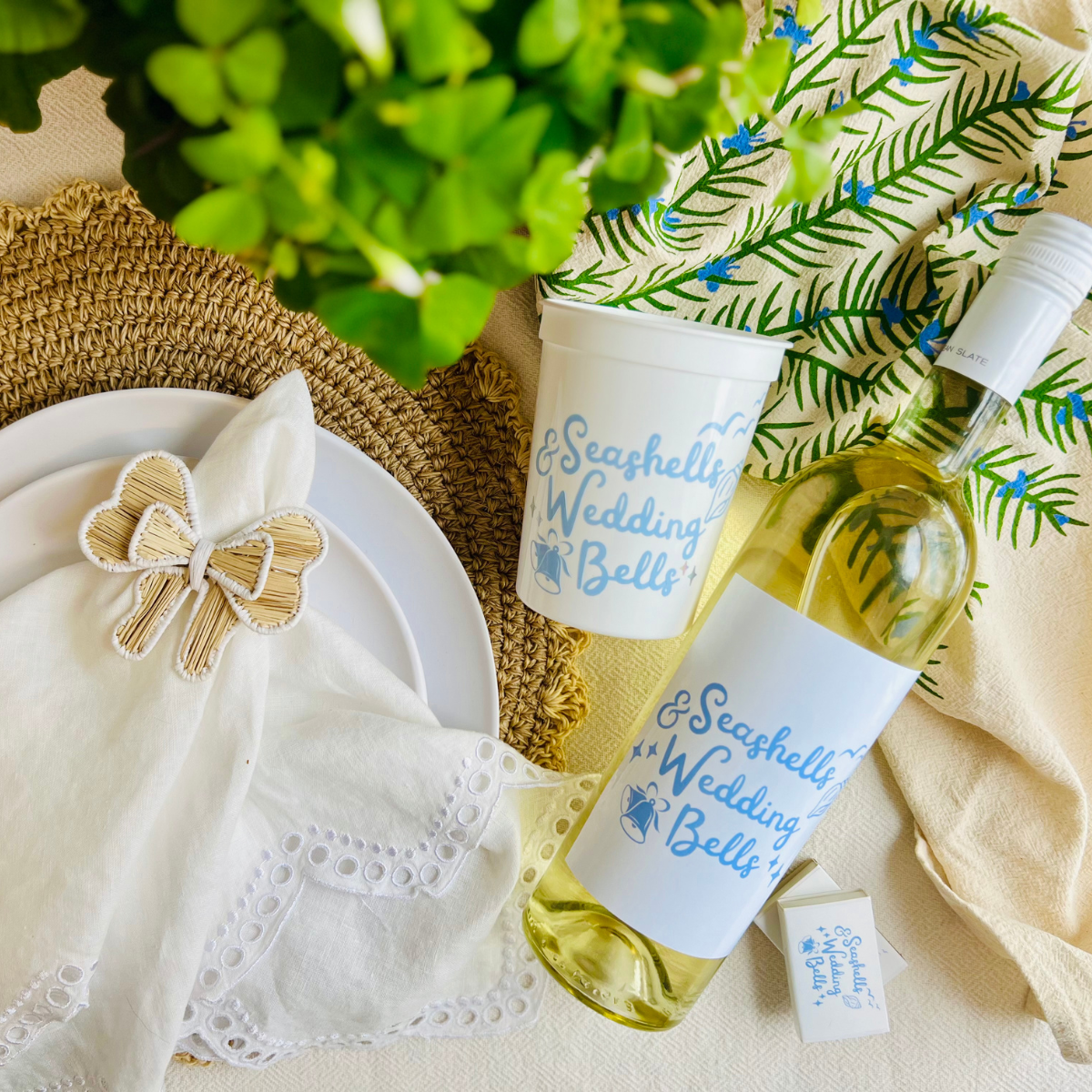 White wine bottle with a "Seashells & Wedding Bells" wine label is shown beside a stadium cup that features a matching design beside a pretty table with greenery and a plate setting.