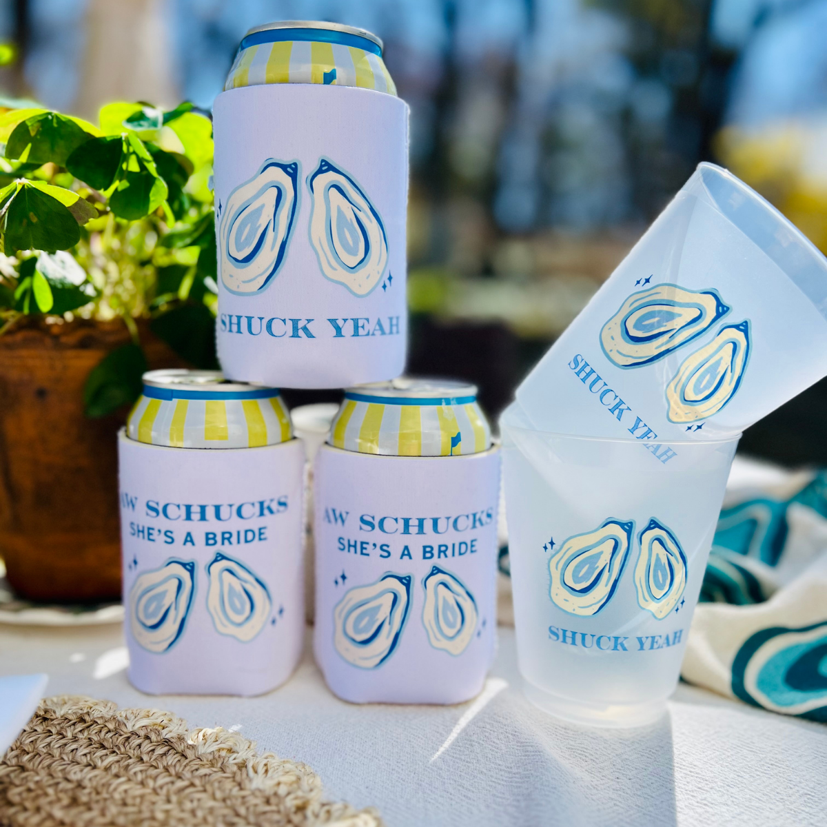 Cans in can cooler sleeves and frost flex cups with 'Shuck Yeah' and 'Aw Shucks, She's A Bride" oyster-inspired design are shown on an outdoor table.
