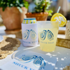 Cocktail with lemon garnish and 'SHUCK YEAH' branded cup on a table with plants in the background.