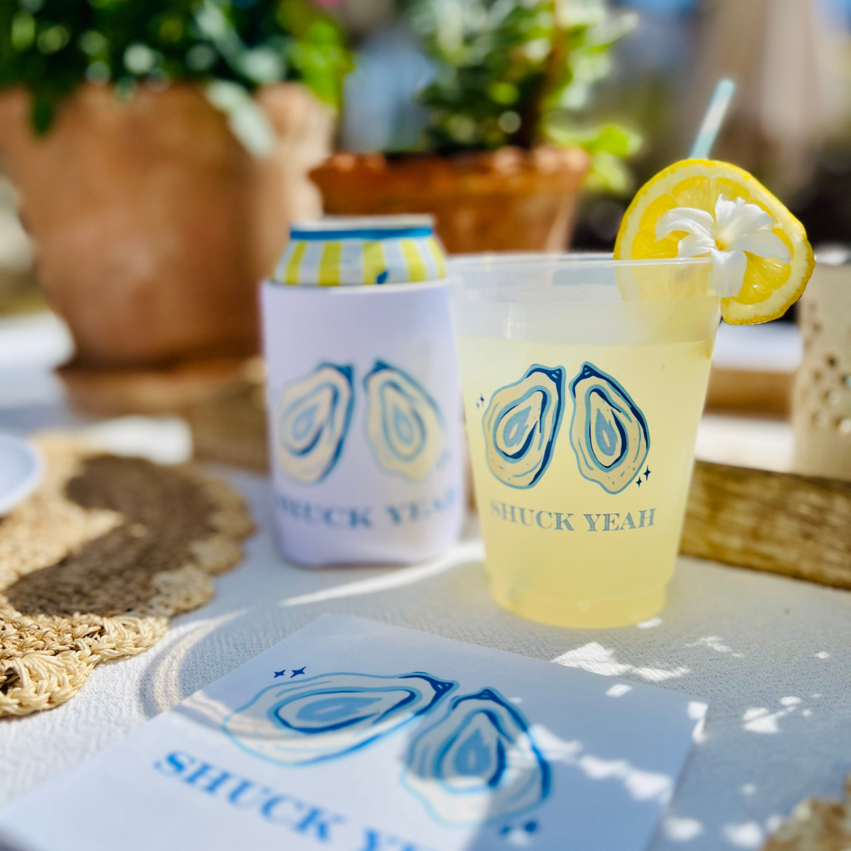 Cocktail with lemon garnish and 'SHUCK YEAH' branded cup on a table with plants in the background.
