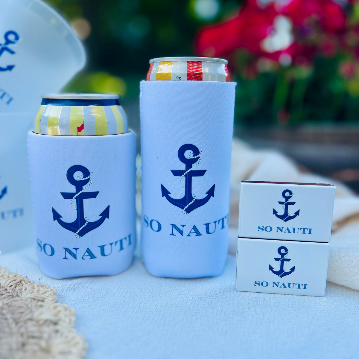 Coozies and matchboxes with anchor design and 'SO Nauti' text on a light blue surface.