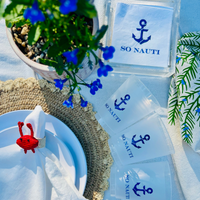 Clear frost flex cups with a blue anchor graphic and blue text that reads "So Nauti" for beach parties, boat parties, and coastal bachelorettes. The cups are shown alongside matching "So Nauti" cocktail napkins, and a plant with blue flowers.