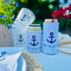 Can coolers with anchor design and 'SO NAUTI' text on a table with flowers and matching cups in the background