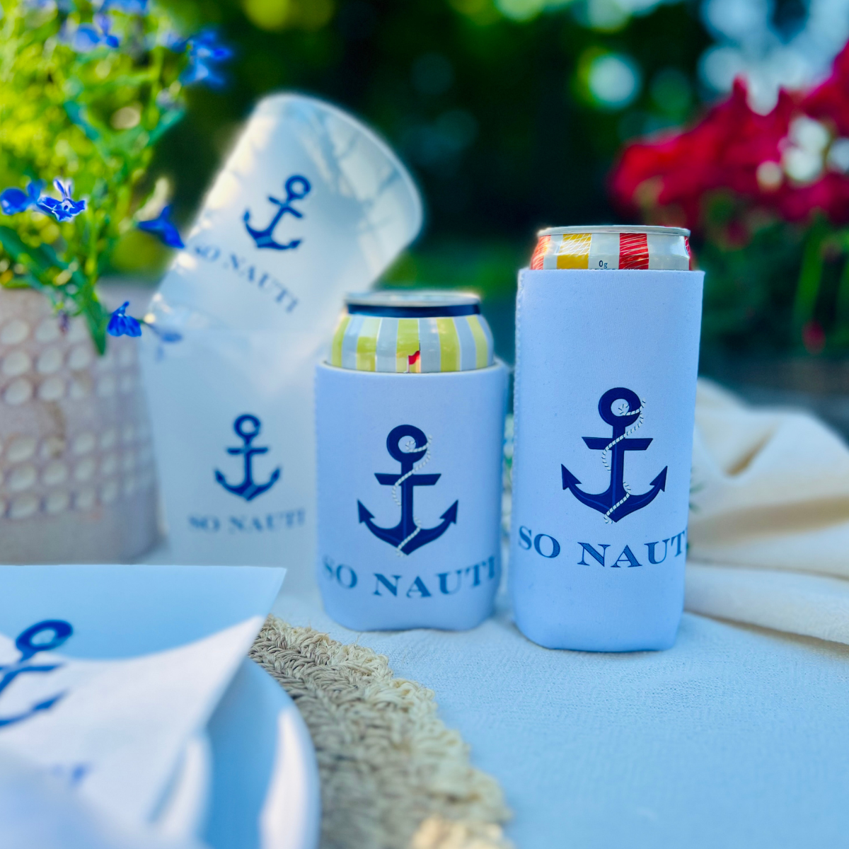 Can coolers with anchor design and 'SO NAUTI' text on a table with flowers and matching cups in the background