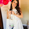 A smiling woman in a white sleeveless dress holds a cream canvas tote bag printed with 'SOON-TO-BE-MRS' and a custom couple's photo. The background features a white banner with pink floral decorations and gold lettering.