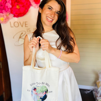 A smiling woman in a white sleeveless dress holds a cream canvas tote bag printed with 'SOON-TO-BE-MRS' and a custom couple's photo. The background features a white banner with pink floral decorations and gold lettering.