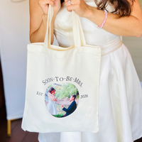 A woman in a white dress holds a cream canvas tote bag with a custom printed circular photo and the text 'SOON-TO-BE-MRS' and 'EST. 2026'.