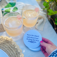 Two cocktails with a garnish on a cocktail pick are shown on a table and a fun coaster that reads "Sorry for what I said when I was docking the boat" is held up with a hand. 