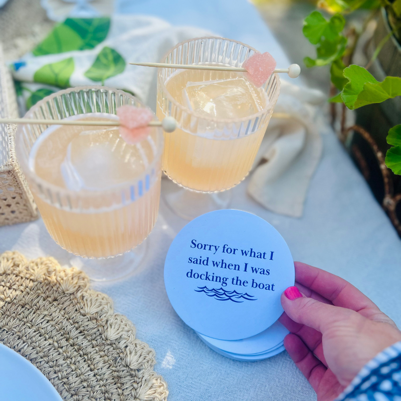 Two cocktails with a garnish on a cocktail pick are shown on a table and a fun coaster that reads "Sorry for what I said when I was docking the boat" is held up with a hand. 