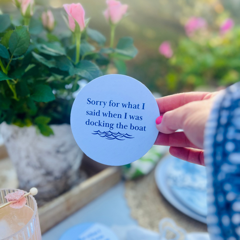 A hand holds up a round white coaster with the text "Sorry for what I said when I was docking the boat." The coaster is shown against a lush garden background and is a perfect accent or gift for a lake house. 