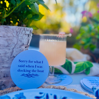 A coasters is shown standing upright and features the text "Sorry for what I said when I was docking the boat." It appears beside a cocktail in the background against greenery in the background.