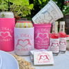 A coordinated set of bachelorette party drink accessories and mini bottles with matching pink and white printed labels reading 'Til Death Do Us Party' and a heart graphic, arranged on a table outdoors with plants in the background.