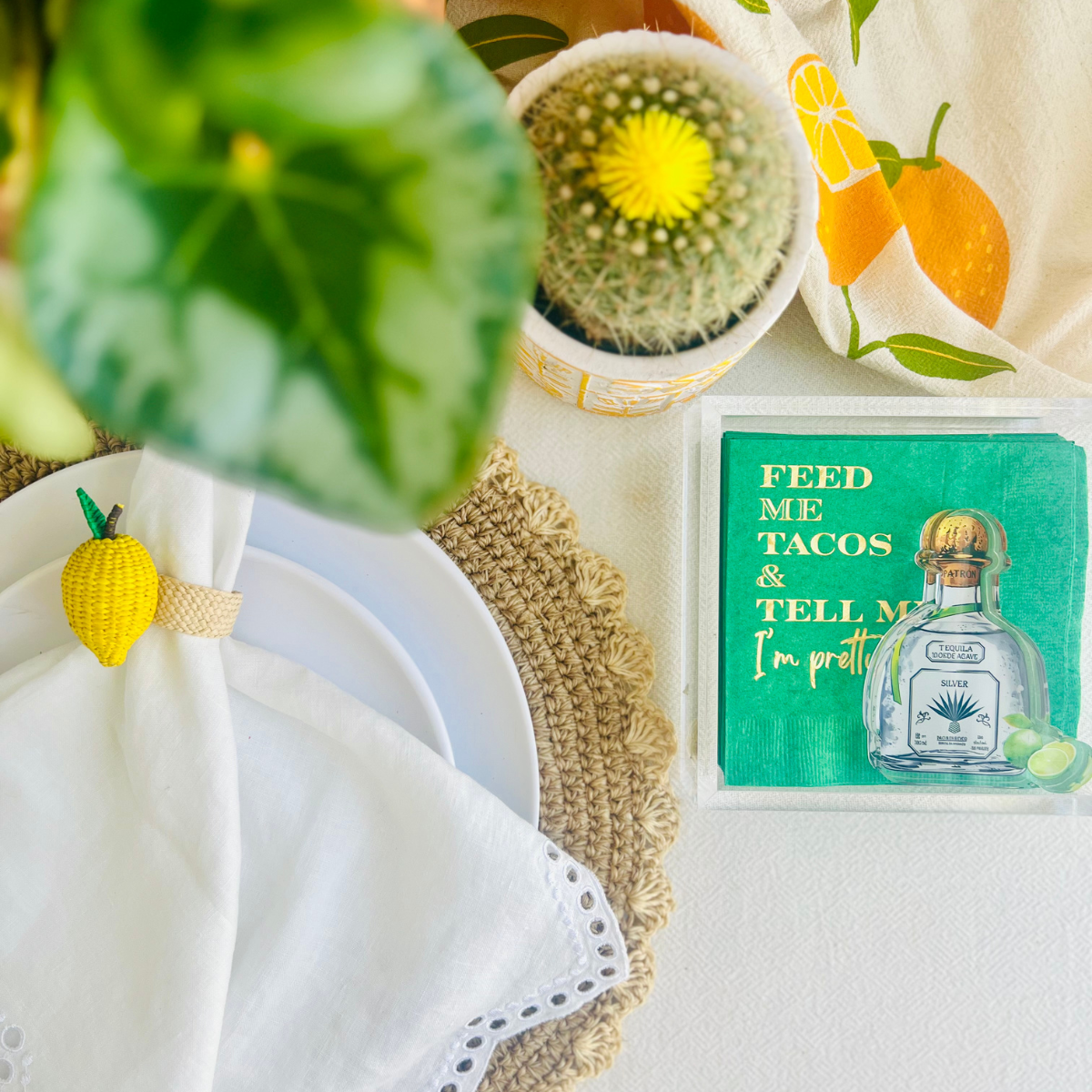 Decorative table setting with an acrylic shaped tequila bottle napkin weight is shown on green  napkin that features text that reads "Feed me tacos and tell me I'm pretty". A lemon napkin ring, and a potted cactus are also shown. 
