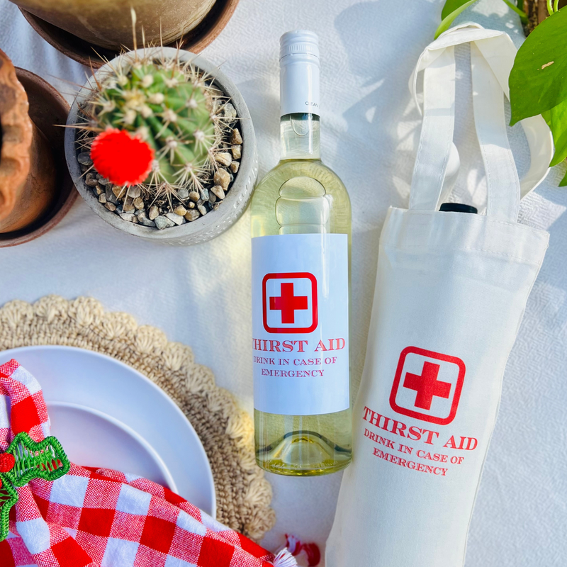 Flat lay of First Aid canvas wine tote and matching emergency wine label on bottle, styled on a table with cactus. 
