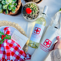 Overhead view of Thirst Aid wine tote beside matching "Drink In Case of Emergency" wine label on bottle, with hand touching tote and funny cocktail napkin reading "They should put more wine in a bottle, so there's enough for two people. 