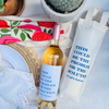 Wine bottle with a label and a white bag with text on a table with a floral cloth and cactus plant. The wine bottle and wine bag feature the same funny quote that reads "This Could Be The Problem Or The Solution - xo Let's Find Out" in blue font. 