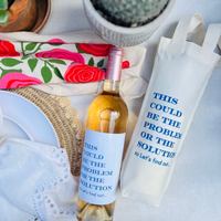 Wine bottle with a label and a white bag with text on a table with a floral cloth and cactus plant. The wine bottle and wine bag feature the same funny quote that reads "This Could Be The Problem Or The Solution - xo Let's Find Out" in blue font. 