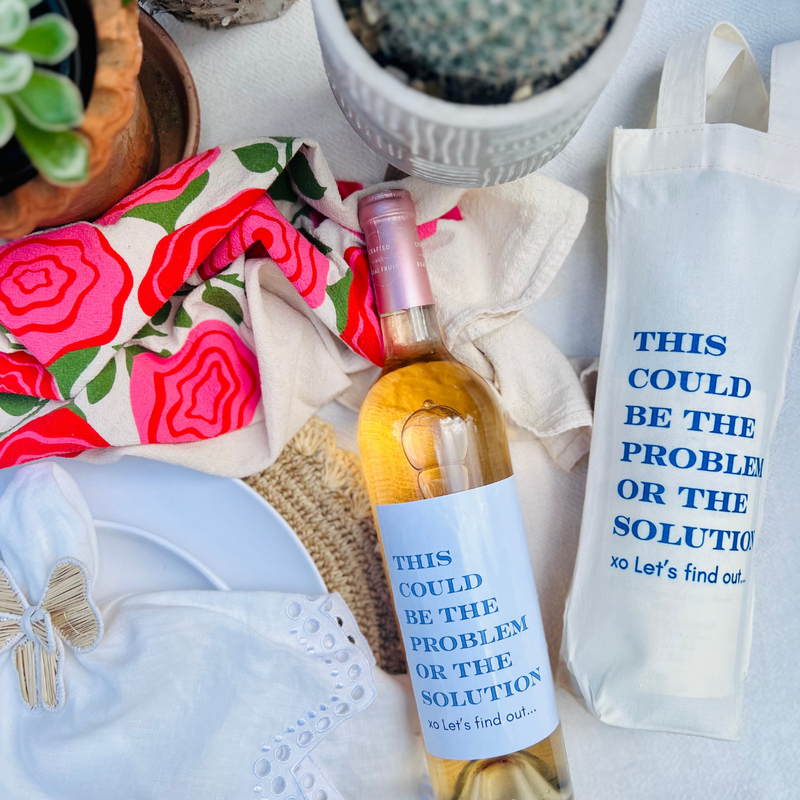 Overhead shot of a wine bottle beside a matching canvas wine bag. Both the bottle and the wine bag feature the fun text "This Could Be The Problem Or The Solution - Let's find out." Perfect for gifting to a hostess, wine lover, or anyone who loves pairing wine with a good laugh. 