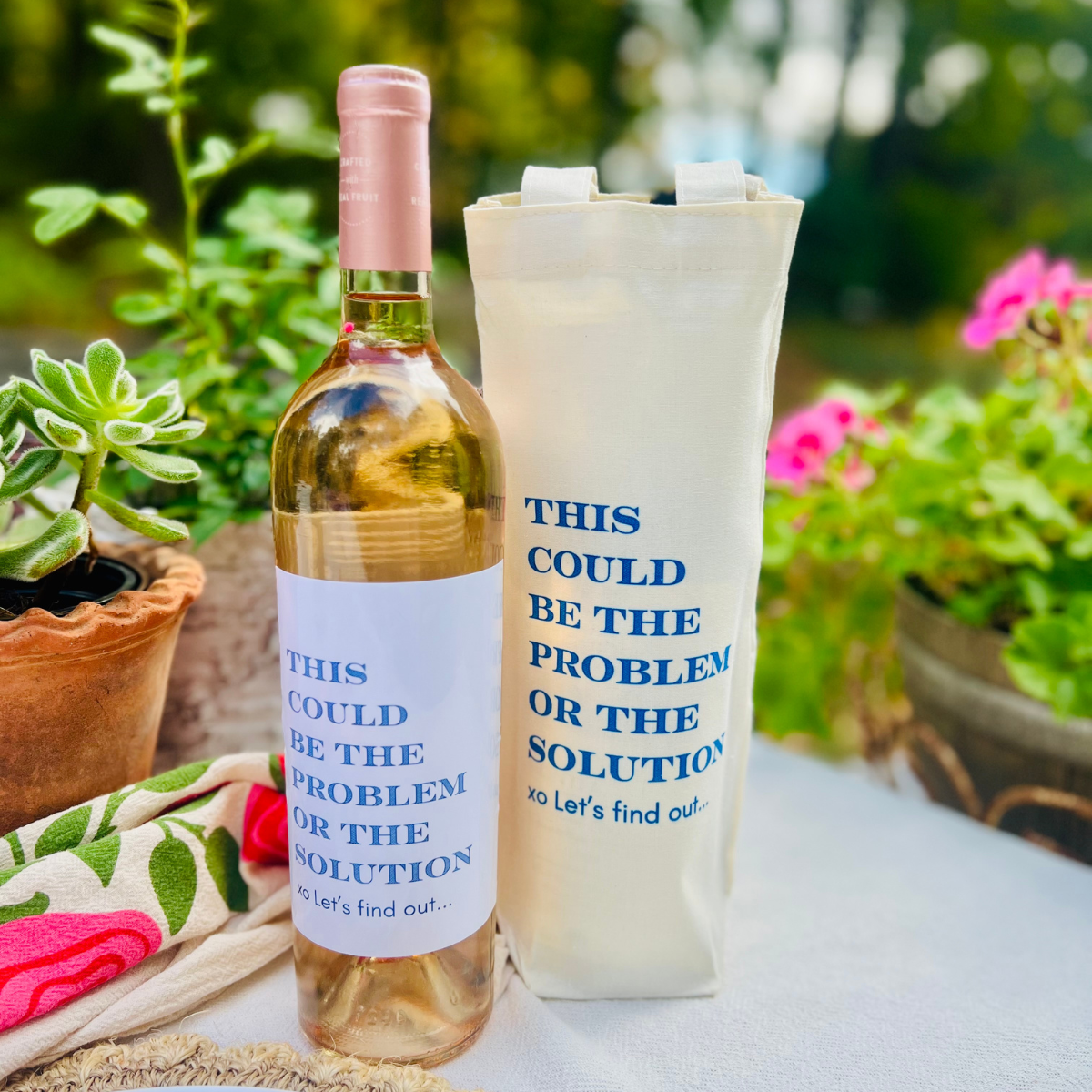 Wine bottle with matching tote bag on a table outdoors. The wine bag and the wine bottle read "This could be the problem or the solution xo Let's find out" in blue text.