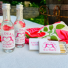 Personalized bachelorette party favors including two mini clear bottles with pink and white labels, a white matchbox, and a matching folded towel with pink roses and green leaves, arranged on a white tablecloth outdoors.