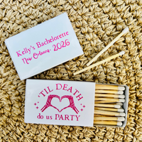 Personalized bachelorette-themed matchbox set on a woven straw background. One closed white matchbox lid shows pink text reading 'Kelly's Bachelorette New Orleans, 2026,' and the open matchbox below shows wooden matches with a white lid printed with 'TIL DEATH do us PARTY' and a pink heart design.