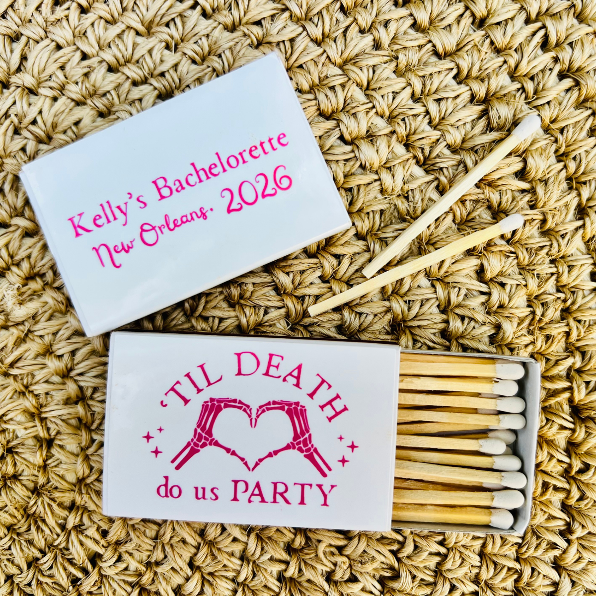 Personalized bachelorette-themed matchbox set on a woven straw background. One closed white matchbox lid shows pink text reading 'Kelly's Bachelorette New Orleans, 2026,' and the open matchbox below shows wooden matches with a white lid printed with 'TIL DEATH do us PARTY' and a pink heart design.