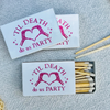 Three white matchboxes with a pink graphic of skeletal hands forming a heart and the text 'TIL DEATH do us PARTY' are arranged on a textured gray fabric background. One matchbox is open, showing wooden matches with white tips.
