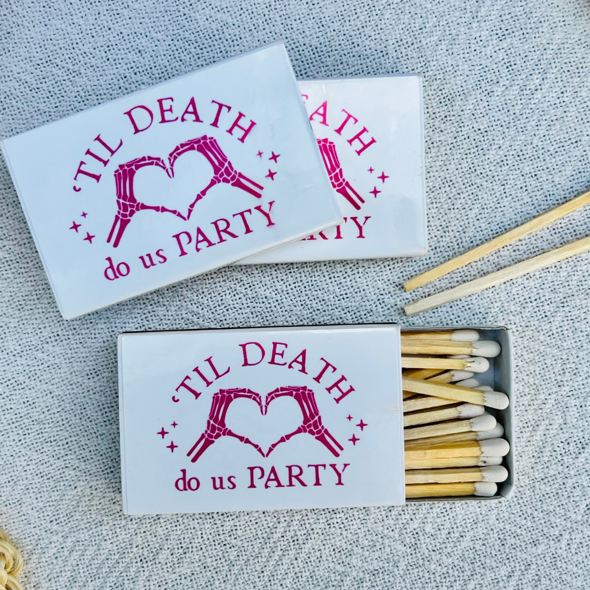 Three white matchboxes with a pink graphic of skeletal hands forming a heart and the text 'TIL DEATH do us PARTY' are arranged on a textured gray fabric background. One matchbox is open, showing wooden matches with white tips.