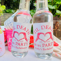Two clear glass bottles with white labels featuring pink skeleton hands forming a heart and the text 'Til DEATH DO US PARTY Kelly's Bachelorette', set outdoors on a table with greenery in the background.