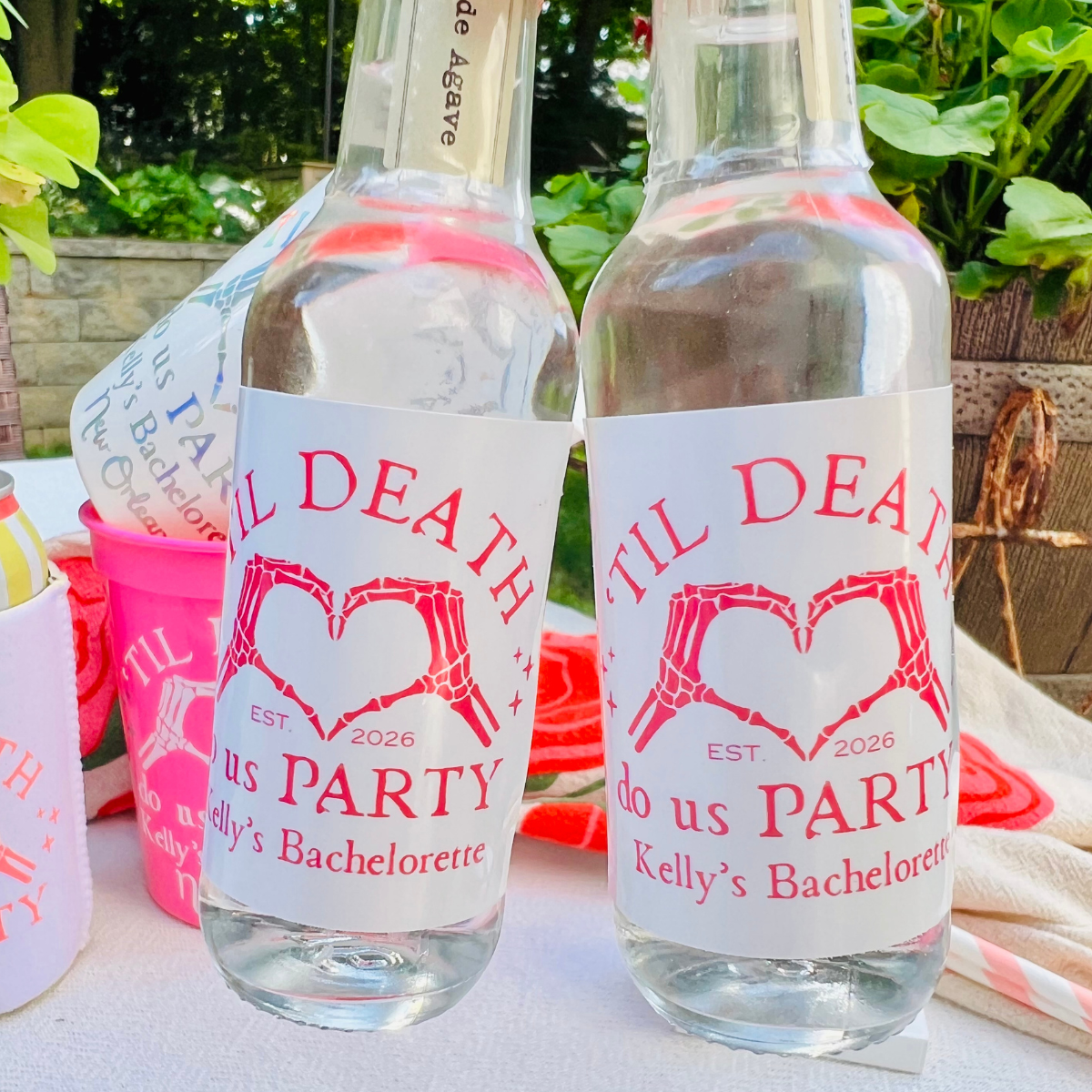 Two clear glass bottles with white labels featuring pink skeleton hands forming a heart and the text 'Til DEATH DO US PARTY Kelly's Bachelorette', set outdoors on a table with greenery in the background.