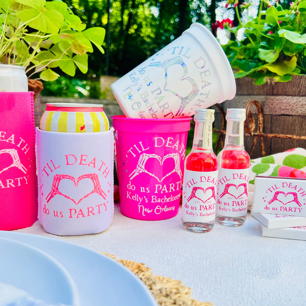 A coordinated party drink set displayed outdoors on a table, including pink and white can coolers, a white cup, two small bottles with tequila, and matching printed koozies and matchboxes. The visible printed text reads 'TIL DEATH do us PARTY' and 'Kelly's Bachelorette New Orleans.