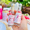 A hand holds two small clear glass bottles with white labels that read “TIL DEATH do us PARTY Kelly’s Bachelorette” in pink text and heart graphics, photographed outdoors with a drink can and party setting blurred in the background.