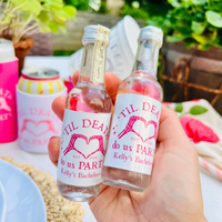 A hand holds two small clear glass bottles with white labels that read “TIL DEATH do us PARTY Kelly’s Bachelorette” in pink text and heart graphics, photographed outdoors with a drink can and party setting blurred in the background.